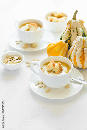 Two cups with fresh pumpkin cream soup decorated cream, seeds and crackers on white table cloth
