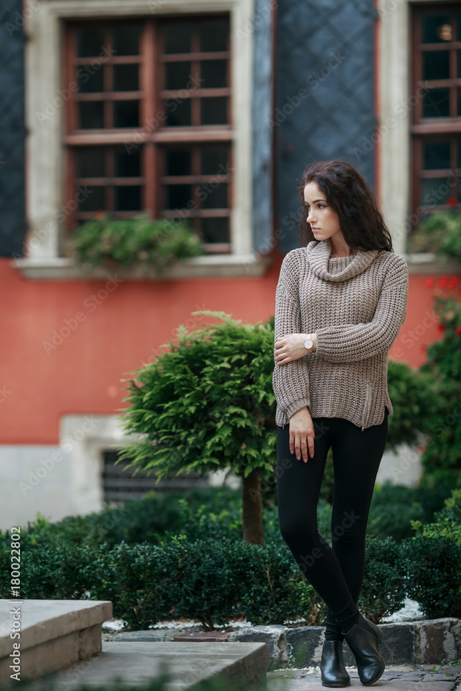 Beautiful young woman dressed in gray sweater and black pants posing in the city on a sunny day. Old town green garden and medieval windows background. Copy space Beautiful young woman dressed in gray sweater and black pants posing in the city on a sunny day. Old town green garden and medieval windows background. Copy space