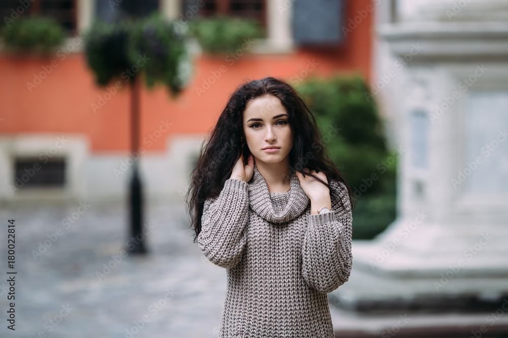 Pretty young woman dressed in gray sweater posing in the mediaeval city on a sunny day. Old town green garden and ancient windows background. Copy space