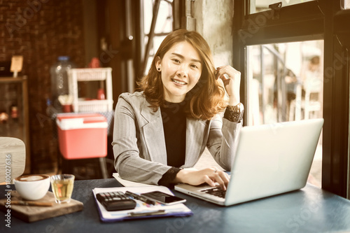 Smiling beautiful Asian girl with black hair working in cafe,Casual woman manager sitting in her office workspace with computer and big bright window and drinking coffee ,look at camera,vintage color