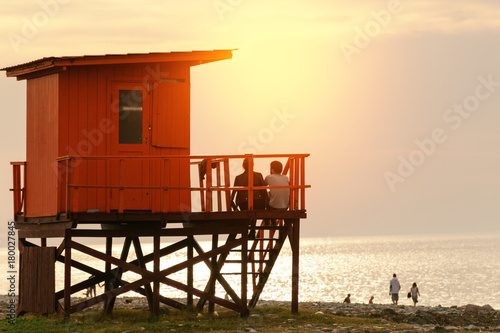 Couple are sitting on the lifeguard tower on the beach, people are walking around. Sunset time. © Lina