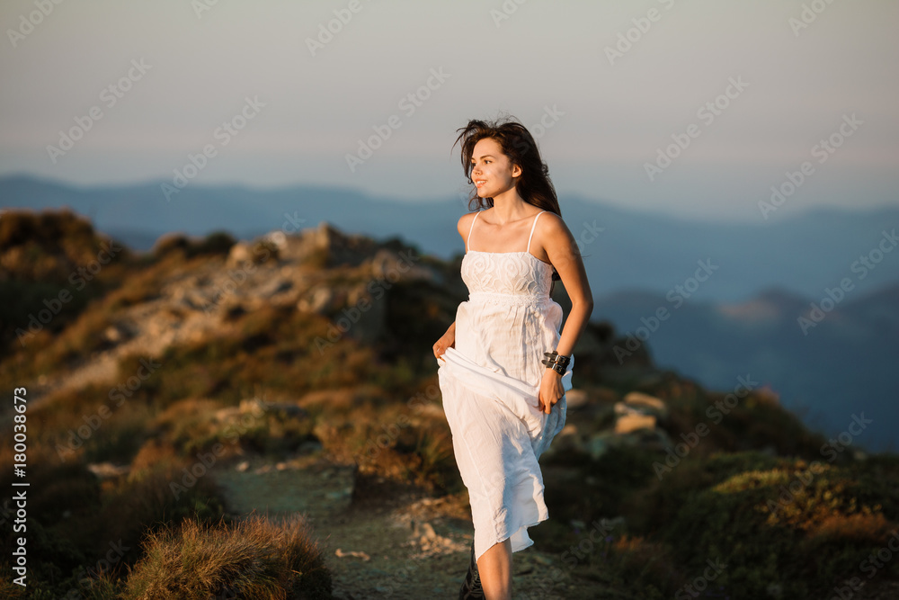 Beautiful woman in white dress running in the mountains. Jumping on mountain peak rock. Beautiful girl looking happy and smiling. Relaxing, feeling alive, breathing fresh air, got freedom from work