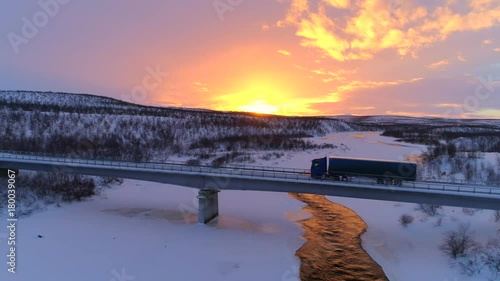 AERIAL: Flying above semi truck driving along scenic countryside highway at sunset. Transporter lorry crossing a bridge above frozen icy river in picturesque snowy landscape at gorgeous golden sunrise
