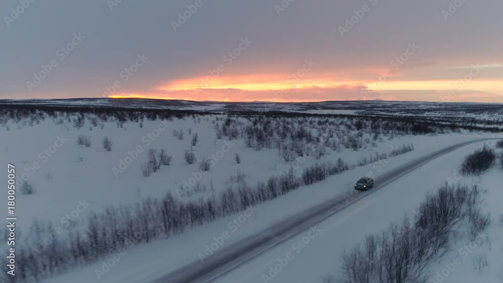 AERIAL: Flying above blue car driving along slippery snowy road in frosty winter backcountry at golden sunrise. People on road trip traveling across snow covered Lapland wilderness at orange sunset AERIAL: Flying above blue car driving along slippery snowy road in frosty winter backcountry at golden sunrise. People on road trip traveling across snow covered Lapland wilderness at orange sunset