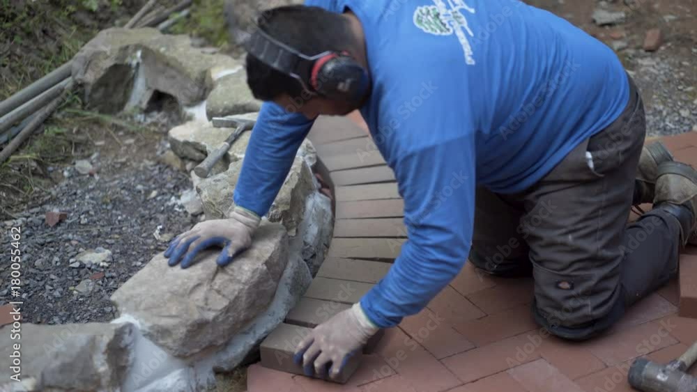 Man with ear protection putting brick pavers in place along an edge of a rock wall. Man with ear protection putting brick pavers in place along an edge of a rock wall.
