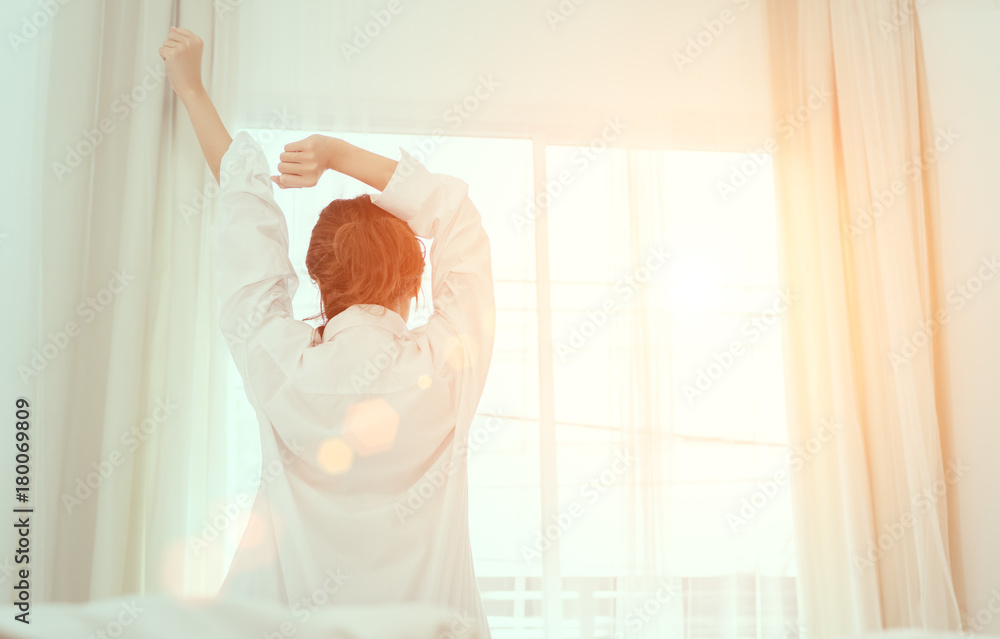 Young woman wake up in the morning and sitting on bed at window door side relaxing in holiday with sunlight, back view - obrazy, fototapety, plakaty 