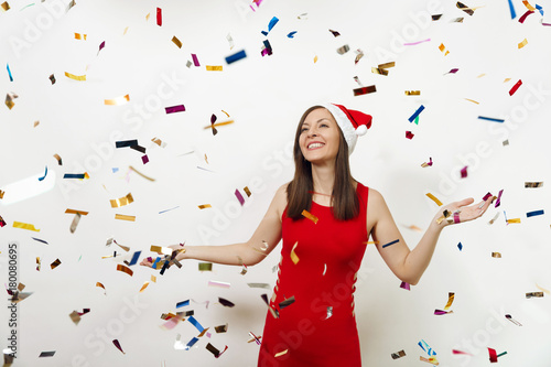 Beautiful caucasian young happy woman with charming smile in red dress and Christmas hat spreading hands on white background and colorful confetti. Santa girl isolated. New Year holiday 2018 concept
