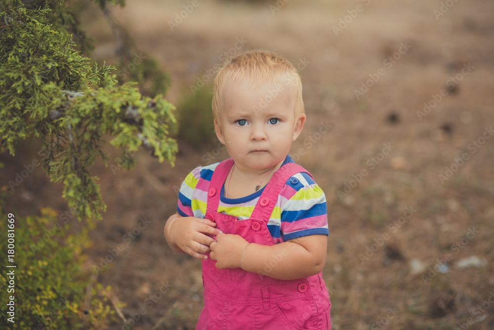 Happy cute baby girl with blond hairs wearing pink jeans dress posing smiling in central park between green tree and stone Happy cute baby girl with blond hairs wearing pink jeans dress posing smiling in central park between green tree and stone