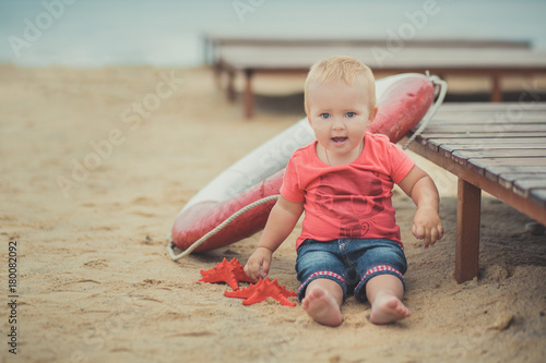 Happy cute baby girl with blond hairs and blue eyes wearing stylish clothes posing smiling on sea side beach on wooden way with safety round and red sea star