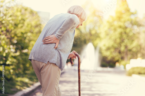 Old age. Sad aged unhappy woman holding a walking stick and walking while feeling pain in the back