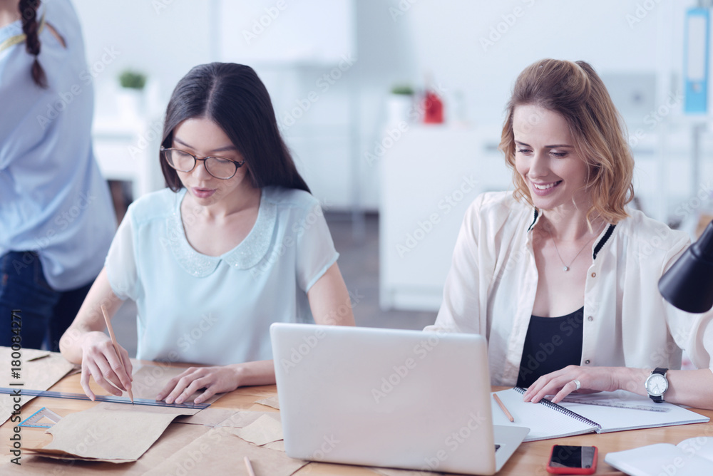 Obraz premium Good beginning. Busy young colleagues working in their modern fashionable atelier while sitting at the table with a laptop and sewing patterns