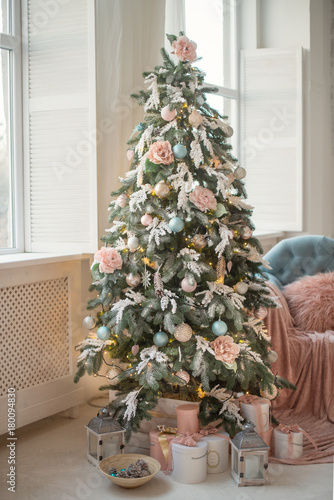 A Christmas tree near the window is decorated with toys and flower in pastel colors. Christmas gifts under the Christmas tree