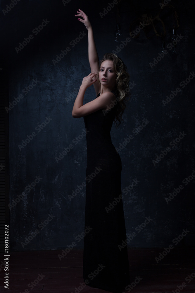 Dark light. A young girl in a beautiful vintage evening long black dress with a chic hairstyle posing with her hands up. Dark light. A young girl in a beautiful vintage evening long black dress with a chic hairstyle posing with her hands up.