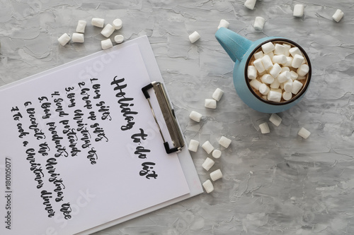 Christmas and New Year cozy holiday composition with to do list, mug with cocoa or chocolate, marshmallow on the gray background. Flat lay, top view.