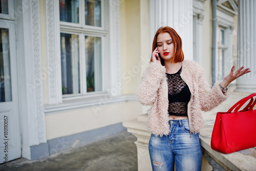 Red haired girl with red handbag posed near vintage house and speaking on mobile phone.