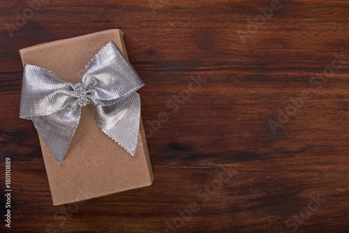 Gift box wrapped in kraft paper with silver ribbon bow on a dark walnut wood. Background top view, close up.