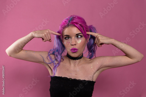 Portrait of funny girl with heavy make up in black top and choker showing screw loose by arms. Studio. Half length. Pink background. photo