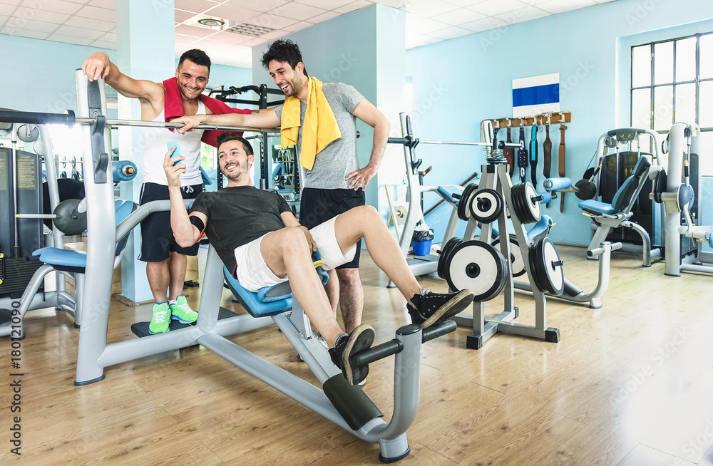 Group of sportive friends using mobile phone at gym fitness club - Happy sporty people in weight room training - Social gathering concept in sport lifestyle context - Main focus on guys interaction Group of sportive friends using mobile phone at gym fitness club - Happy sporty people in weight room training - Social gathering concept in sport lifestyle context - Main focus on guys interaction