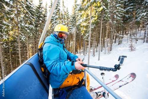 Male skier taking selfies with action camera on selfie stick while riding up on ski lift in the mountains at the winter resort