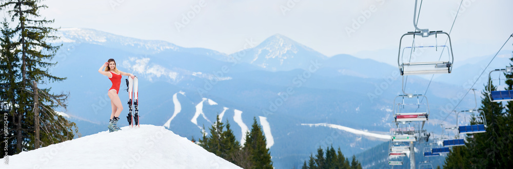 Fototapeta premium Panoramic view of winter ski resort Bukovel. Beautiful woman skier wearing red swimsuit, posing with skis on the top of the snowy slope. Mountains, forests, ski slopes and ski-lift on the background