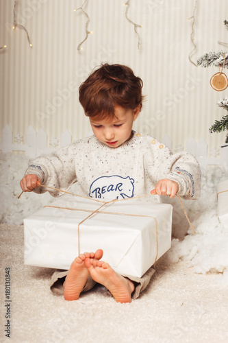 Cute little boy is sitting with a gift near the Christmas tree. The child is happy with New Year's gift. Caucasian blonde toddler boy