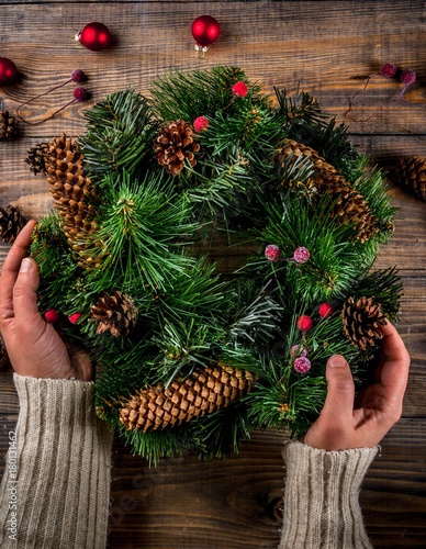 Preparation for xmas holidays. Woman decorating christmas green wreath with pine cones and red winter berries and christmas tree balls  wooden background  top view copy space  female hands in pictures