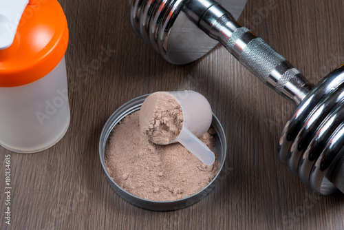 Whey protein in a measuring bucket with a metal dumbbell and shaker on a wooden background