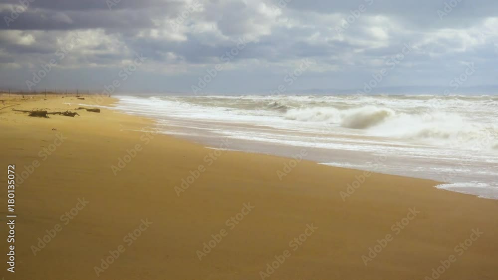 A sandy beach with a fence in the distance and a stormy sea with waves under a cloudy sky, a shadow from a cloud rushes to the beach