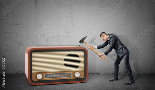 A tiny businessman on concrete background tries to destroy a large retro radio set with a broken hammer.
