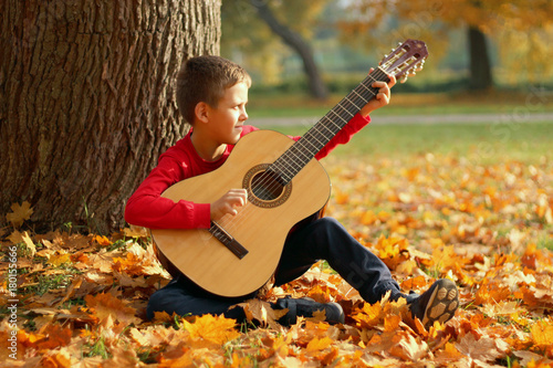 A boy in a red sweater plays a guitar near a tree