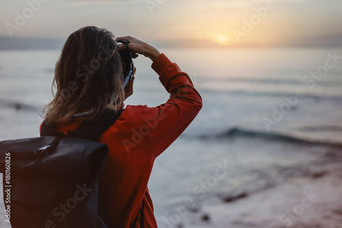 Hipster hiker tourist with backpack taking photo of seascape sunset on camera on background sea, photographer enjoying ocean horizon, blurred panoramic sunrise, traveler relax holiday concept trip