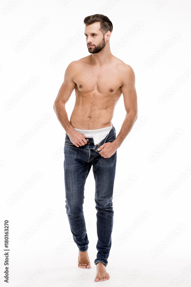 Casual and gorgeous. Full length of handsome shirtless young man taking off his jeans and looking away while standing against white background.