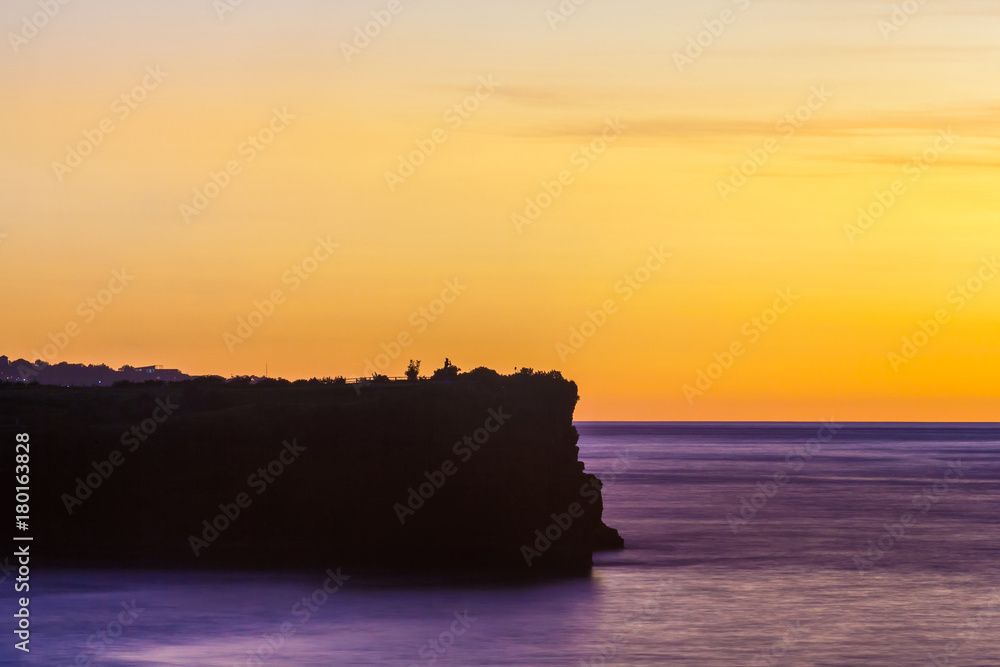 Silhouette of a cliff against the background of an orange sunset. Sea waves break on the rocks. Sunset on Jimbaran, South Kuta, Bali, Indonesia.