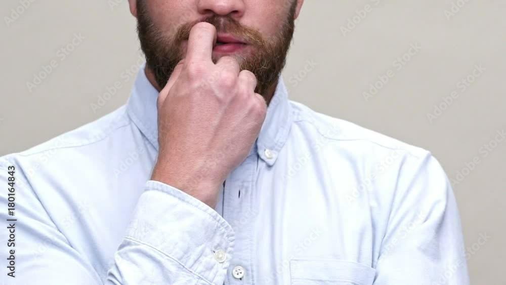 Cropped close up view of bearded man in shirt bites his finger over gray background Cropped close up view of bearded man in shirt bites his finger over gray background
