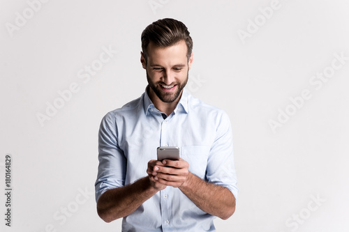 Great news! Handsome young man using his phone with smile while standing against white background.