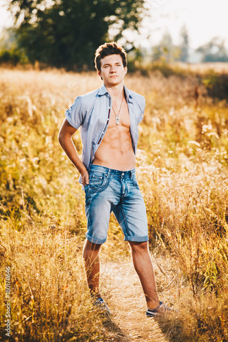 a handsome young sexy man with a strong muscular torso in an unbuttoned shirt is standing on a meadow in nature outside city © Elizaveta