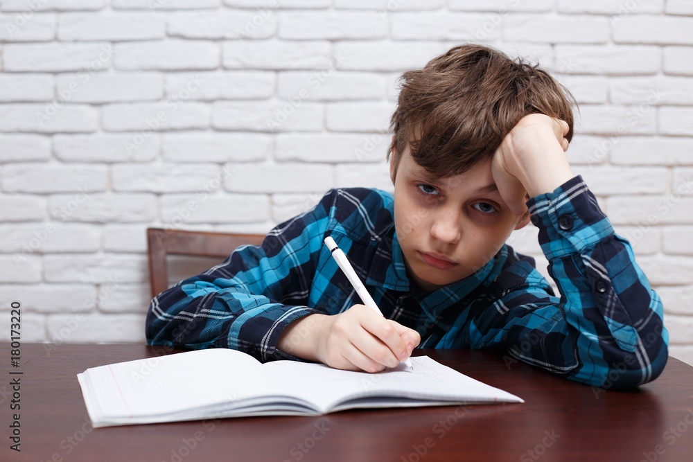Bored schoolboy writing down a task into a notebook. Studying difficulties, elementary school, school life, education, homework, school concept Bored schoolboy writing down a task into a notebook. Studying difficulties, elementary school, school life, education, homework, school concept