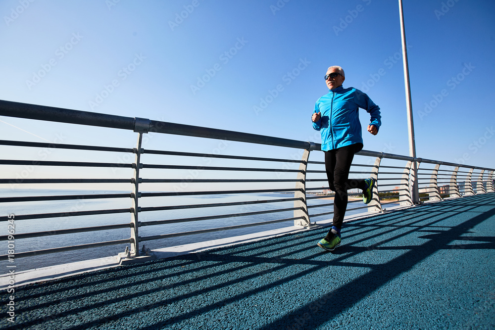 Contemporary pensioner running in the morning on urban bridge by riverside