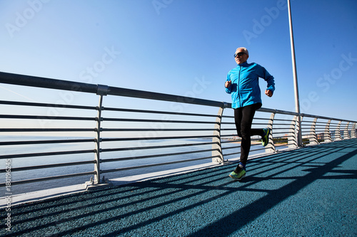 Contemporary pensioner running in the morning on urban bridge by riverside