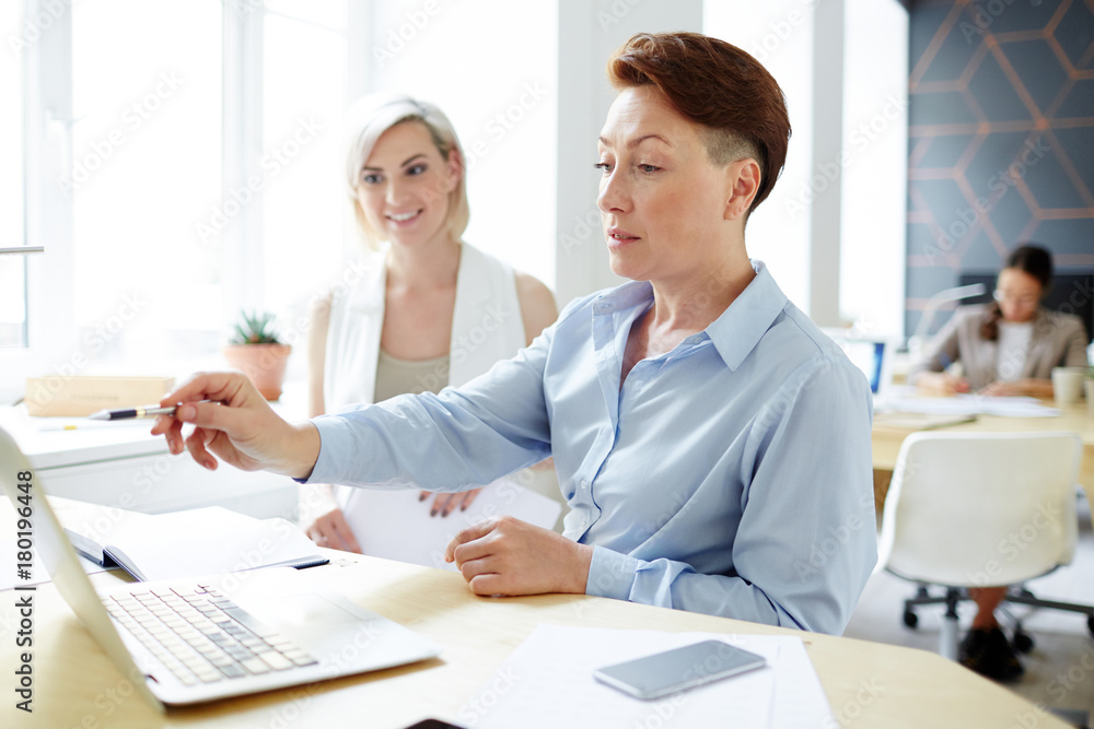 Experienced analyst pointing at laptop display while explaining her subordinate how to work with data Experienced analyst pointing at laptop display while explaining her subordinate how to work with data