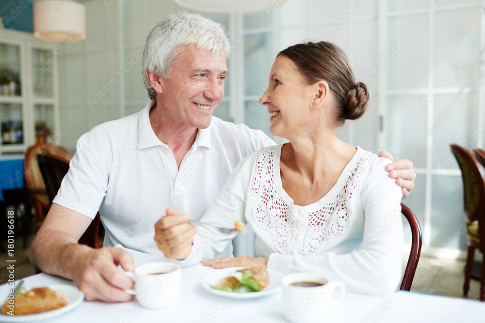 Mature affectionate couple sitting in cafe, eating dessert with tea or coffee and having talk