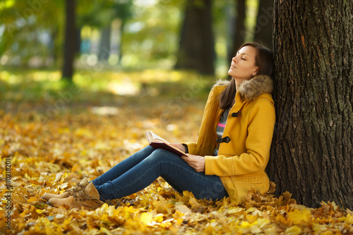 A beautiful happy smiling brown-haired woman in yellow coat and jeans sitting under the maple tree with a red book in fall city park on a warm day. Autumn golden leaves. Reading concept