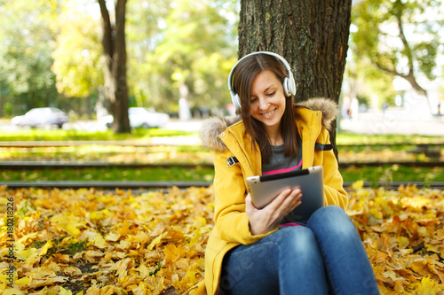 A brown-haired smiling woman in yellow coat and jeans sitting and listening to music under a tree with a tablet in her hands and headphones in fall city park on a warm day. Autumn golden leaves.