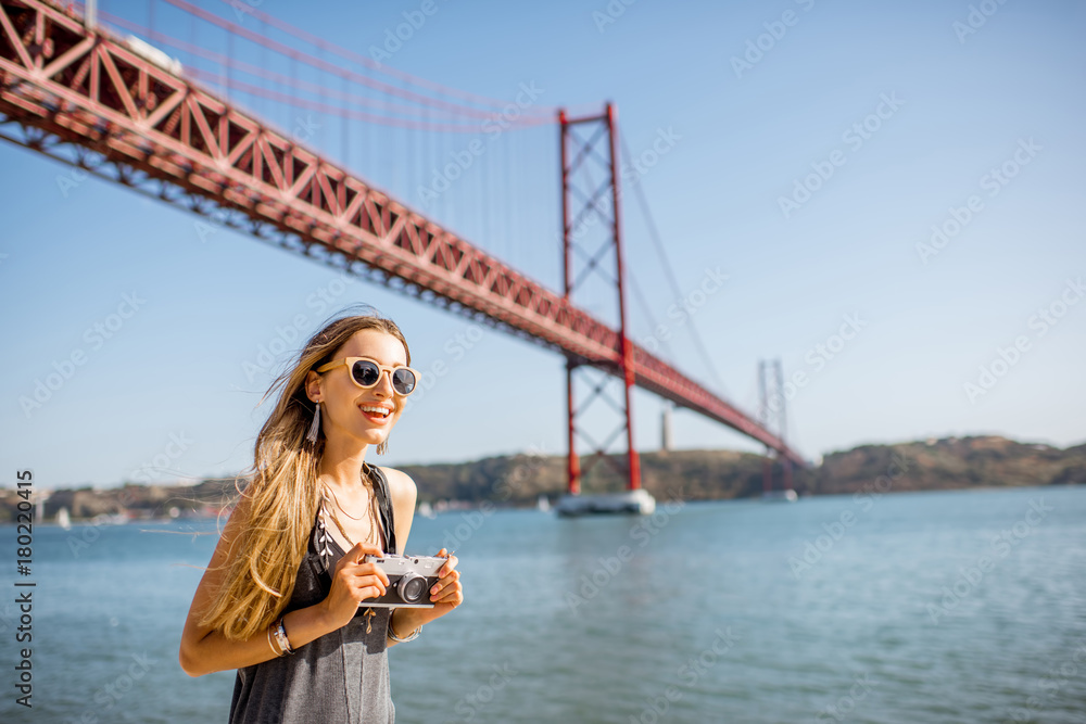 Portrait of a young woman tourist with photo camera standing in front of the famous iron bridge traveling in Lisbon city, Portugal Portrait of a young woman tourist with photo camera standing in front of the famous iron bridge traveling in Lisbon city, Portugal