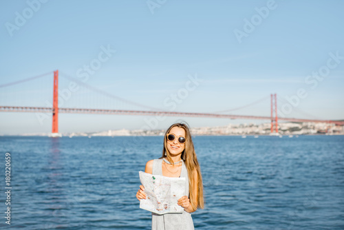 Portrait of a woman tourist standing with paper map on the riverside with beautiful iron bridge on the background in Lisbon city, Porugal