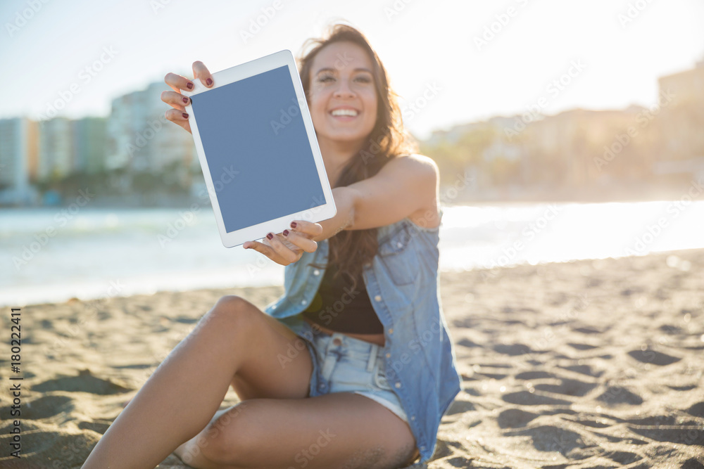Young happy woman sitting on beach showing tablet
