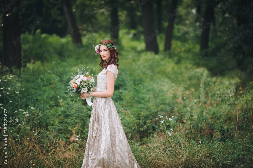An elegant bride with a wreath on her head and a light cream wedding dress is standing in a summer forest. Walk on the nature.