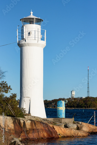 Small white lighthouse on the Karlskrona (Sweden) archipelago island of Stumholmen with coastal landscape in background. Water tower and radio antenna on island behind lighthouse. photo