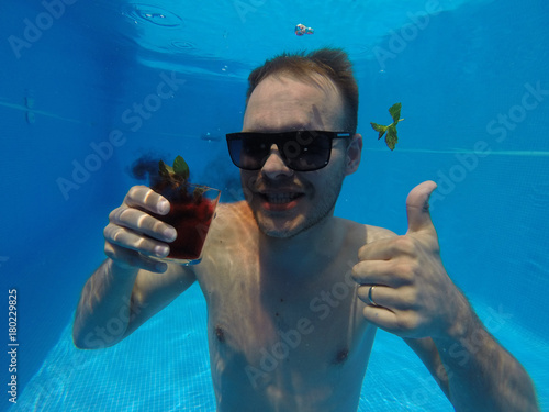one unshaven man drinks an alcohol cocktail under the water in a pool with sunglasses in the summer, shows thumb