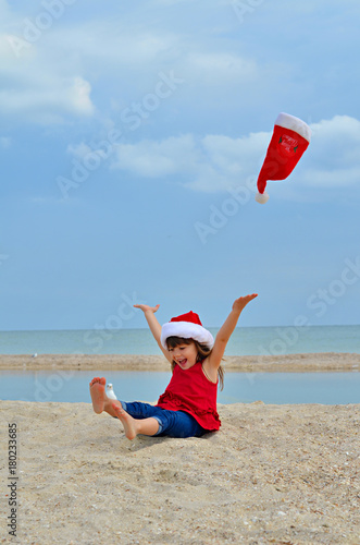Fototapeta Naklejka Na Ścianę i Meble -  Cute little girl celebrating Christmas and New Year holidays on the beach, throwing up Santa hat